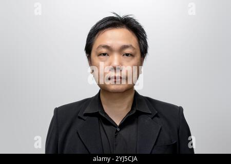 Uomo asiatico di mezza età, vestito con un elegante abito nero e una camicia, posa su uno sfondo bianco-grigio neutro. Foto Stock