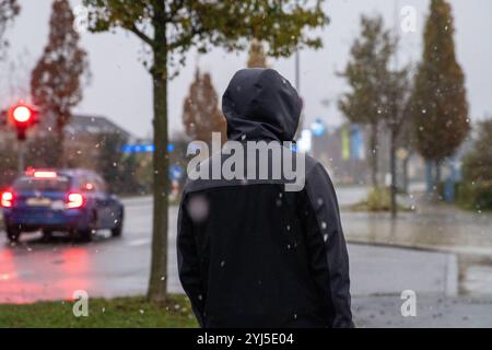 Langweid, Baviera, Germania - 13 novembre 2024: Immagine simbolica dei cambiamenti climatici e delle nevicate in Baviera. Un uomo con un cappuccio e una giacca invernale cammina vicino a una strada con il tempo nuvoloso *** Symbolbild Wetterumschwung und Schneefall a Bayern. Ein Mann mit Kaputze und Winterjacke läuft bei trübem Wetter neben einer Straße spazieren Foto Stock