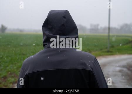 Langweid, Baviera, Germania - 13 novembre 2024: Immagine simbolica dei cambiamenti climatici e delle nevicate in Baviera. Un uomo con un cappuccio e una giacca invernale cammina nel tempo nuvoloso *** Symbolbild Wetterumschwung und Schneefall a Bayern. Ein Mann mit Kaputze und Winterjacke läuft bei trübem Wetter spazieren Foto Stock