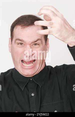 Uomo di mezza età, con capelli scuri su sfondo bianco, fa il viso insolito, piange, urla, starnuti, soffocamento, espressione molto espressiva Foto Stock