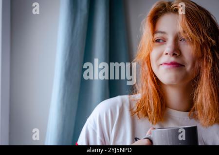 Un'elegante ragazza rossa di generazione Z viene vista sorseggiare una bevanda calda mentre si trova accanto a una finestra, abbracciando il calore e la serenità in una candida Foto Stock