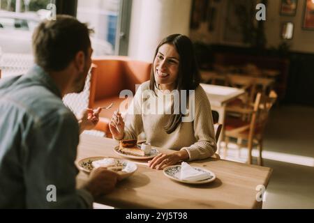 In un caffè caldo e invitante, una coppia condivide risate e deliziose prelibatezze, assaporando caffè e pasticcini mentre si crogiola al sole del pomeriggio. Foto Stock