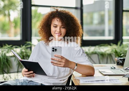 La donna si rilassa mentre legge un libro e controlla il suo telefono in un ambiente d'ufficio vivace Foto Stock