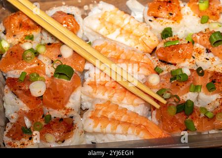 Vista ravvicinata di un contenitore di plastica riempito di sushi, con salmone, gamberi e riso bianco ricoperto di cipolle verdi e peperoncino rosso. Legno Foto Stock