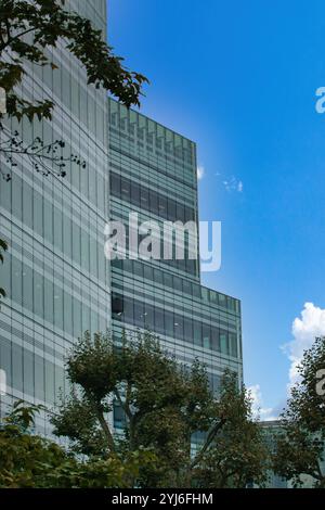 Un moderno edificio in vetro con una facciata a griglia, parzialmente oscurata dagli alberi e un cielo blu con nuvole. Foto Stock