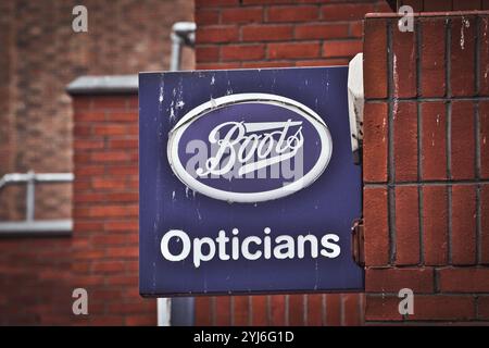 Un primo piano di un cartello blu con il testo bianco "Boots Opticians". Il cartello e' montato su un muro di mattoni con un edificio in mattoni sfocati nella b Foto Stock