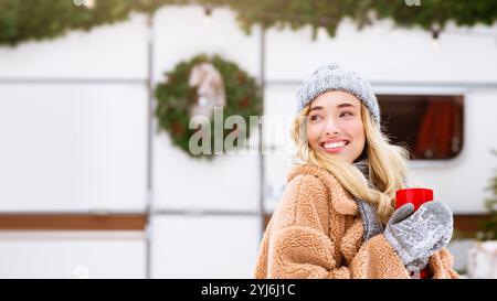 Una giovane donna sorridente sorridente sorregge una bevanda calda mentre indossa un intimo abbigliamento invernale. Si trova vicino alle decorazioni natalizie, catturando l'essenza Foto Stock