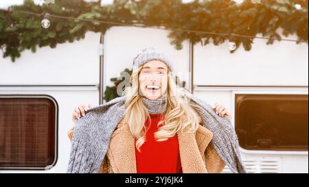 Una giovane donna gioiosa con i capelli biondi indossa un maglione caldo e una sciarpa, che posa allegramente di fronte a un camper decorato durante le festività invernali, capt Foto Stock