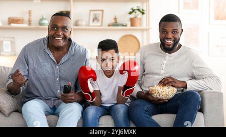 Padre Nero, figlio e nonno entusiasti che guardano la partita di pugilato in TV e Cheering, mangiano popcorn e si rilassano a casa Foto Stock