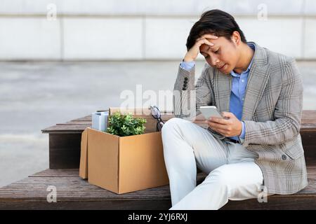 Un uomo asiatico senza lavoro con un blazer grigio e pantaloni bianchi siede su una panchina di legno. Ha i suoi effetti personali in una scatola di cartone accanto a lui. Guarda in basso la sua Foto Stock