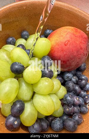 primo piano di frutta fresca, mirtilli, pesca e uva, in un recipiente sotto l'acqua corrente mentre si lava la frutta. Foto Stock