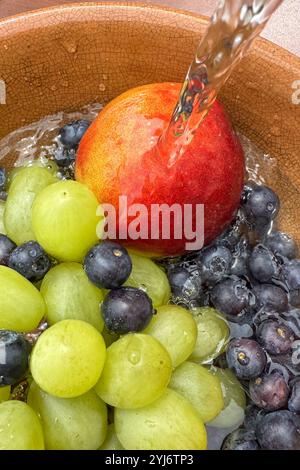 primo piano di frutta fresca, mirtilli, pesca e uva, in un recipiente sotto l'acqua corrente mentre si lava la frutta. Foto Stock