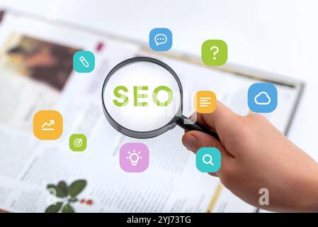 Mano che tiene in mano una lente d'ingrandimento per esaminare un libro con testo SEO, icone concettuali per idee, cloud, ricerca, destinazione e testo che fluttuano Foto Stock