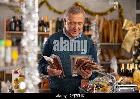 Un uomo maturo con un cestino di alimentari acquista salsiccia secca e prosciutto al supermercato Foto Stock