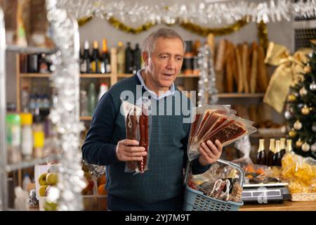 Un uomo maturo con un cestino di alimentari acquista salsiccia secca e prosciutto al supermercato Foto Stock