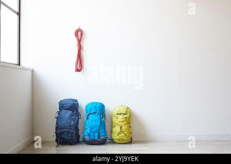 Corda da arrampicata appesa alla parete bianca accanto alla finestra e tre zaini di diverse dimensioni collocati sotto di essa Foto Stock