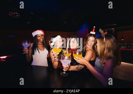 Un gruppo di amici festeggia con gioia le festività natalizie con un drink in un night club, indossando cappelli di Babbo Natale e sorridendo. Foto Stock