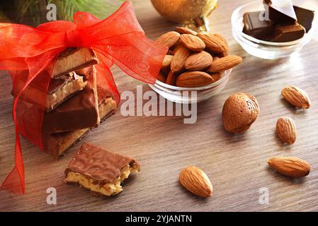 Torrone ricoperto di cioccolato con pezzi impilati legati con nastro rosso su una panca di legno con mandorle naturali e porzioni di cioccolatini e decorazioni natalizie Foto Stock
