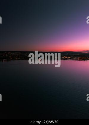 Tramonto spettacolare su un tranquillo paesaggio urbano, che riflette vivaci sfumature rosa e viola sul tranquillo lungomare, con le luci della città che brillano al tramonto. Foto Stock