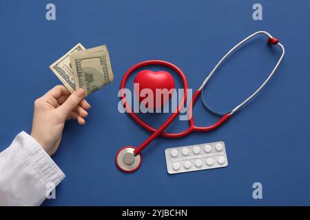 Medico donna che tiene i soldi vicino al cuore, pillole e stetoscopio su sfondo blu. Concetto di assicurazione sanitaria Foto Stock