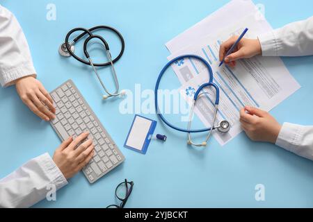 Mani del medico scrivere su assicurazione medica e digitare su tastiera del computer vicino allo stetoscopio su sfondo blu Foto Stock