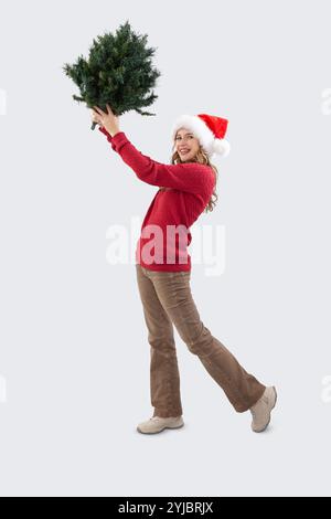 Buon Natale, felice donna sorridente che tiene in mano la cima dell'albero per realizzare e decorare l'albero di Natale, guardando la macchina fotografica che indossa il cappello di Babbo Natale e il rosso Foto Stock