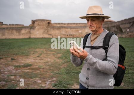 Un turista anziano che esplora l'antica fortezza con lo smartphone durante il viaggio, ammirando l'architettura storica e catturando i ricordi attraverso il moderno Foto Stock