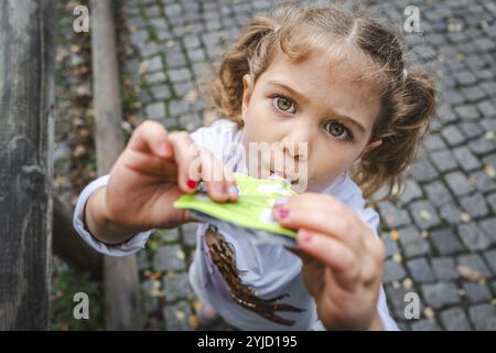 Una giovane ragazza con le trecce mangia uno spuntino in una strada acciottolata e guarda curiosamente, Stoccarda, Baden-Wuerttmeberg, Germania, Europa Foto Stock