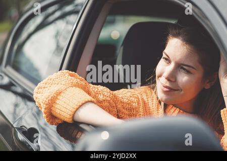 Bella giovane felice sorridente donna caucasica che guida in macchina, indossa occhiali da sole e un maglione arancione. Foto Stock