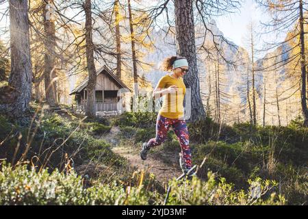 Donna gioiosa che corre nella foresta autunnale, indossa una camicia arancione e leggings colorati. Donna che salta in aria mentre cammina attraverso le fores Foto Stock