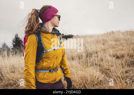 Escursionista caucasica con giacca gialla e zaino rosa che fa trekking in montagna in un giorno d'autunno, indossando guanti e fascia Foto Stock