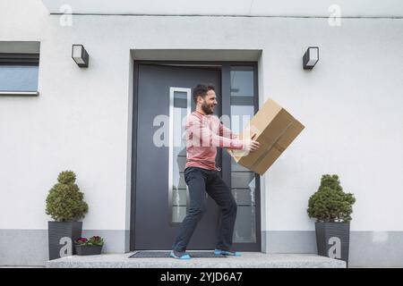 Entusiasta uomo caucasico in piedi davanti alla porta d'ingresso con una grande scatola di cartone in mano che è appena arrivata nella posta. Uomo che riceve un pacchetto eccitante Foto Stock