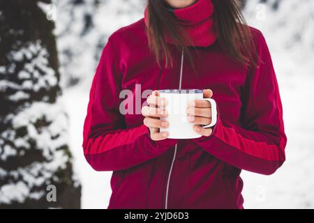 Bella giovane donna caucasica con i capelli castani, che indossa una giacca rossa e un cappello fuori nella foresta quando nevica. Donna che cammina intorno a una neve Foto Stock