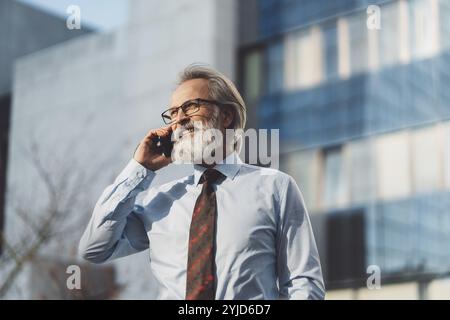 Uomo d'affari anziano con capelli grigi e barba che parla al telefono fuori in una giornata di sole. CEO alla chiamata fuori Foto Stock