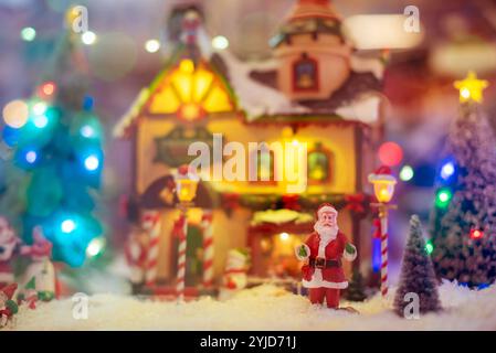 Statuetta di Babbo Natale in miniatura in piedi sulla neve di fronte a un villaggio di natale illuminato con alberi di natale e luci sfocate Foto Stock