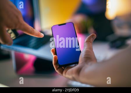 smartphone da studio per l'ufficio mockup a schermo vuoto colori pastello al neon puntando le dita sullo sfondo del laptop Foto Stock