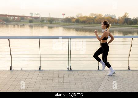 Un determinato corridore procede con sicurezza lungo un sentiero lungo il fiume, abbracciando l'aria frizzante del mattino, con lo skyline della città e gli alberi autunnali che offrono un'immagine Foto Stock