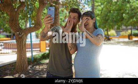 Coppia che fa una videochiamata nel parco mentre si gode una giornata di sole, con la donna incinta che mostra la pancia ed entrambi esprimono emozioni Foto Stock