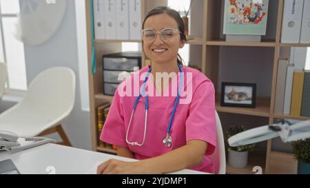 Giovane lavoratrice sanitaria che indossa scrub rosa, seduta in un ufficio della clinica, sorridendo alla telecamera. Foto Stock