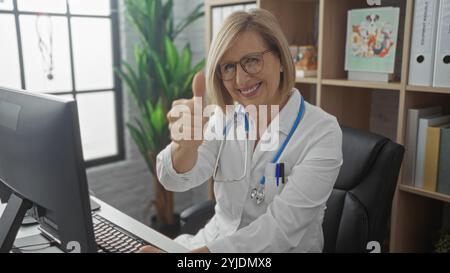 Una donna bionda felice che mostra i pollici mentre si siede in una stazione di lavoro clinica con attrezzature moderne e scaffali sullo sfondo. Foto Stock