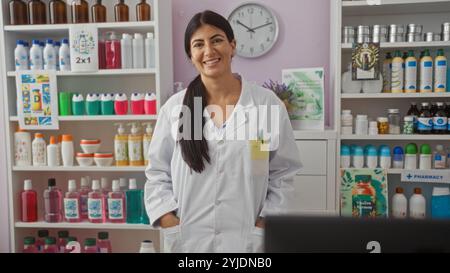 Una giovane donna sorridente farmacista si trova in una farmacia, circondata da scaffali ben organizzati di farmaci e prodotti per la salute. Foto Stock