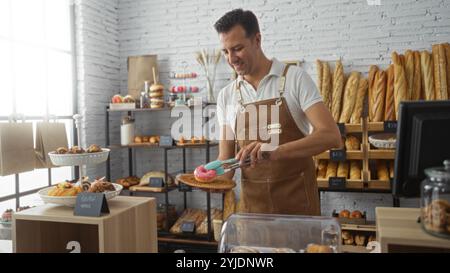 Uomo ispanico che lavora in una panetteria, tenendo una ciambella rosa con pinze, circondato da vari dolci in un ambiente da forno interno Foto Stock