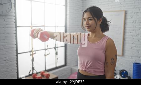 Una giovane donna in palestra solleva un manubrio rosa sorridendo, indossando un top rosa e mostrando tatuaggi sulle braccia, con attrezzature da palestra nel bac Foto Stock