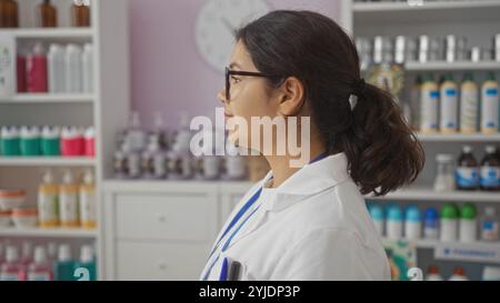 Una donna giovane, attraente, ispanica, bruna con un cappotto bianco si trova all'interno di una farmacia o farmacia ben fornita con vari prodotti e bottiglie Foto Stock