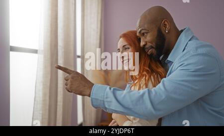 Coppia felice che si gode un momento tenero insieme a casa mentre guarda fuori da una finestra in un accogliente soggiorno Foto Stock