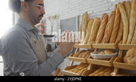 Giovane ispanico che indossa occhiali e grembiule che lavora in una panetteria con scaffali per il pane sullo sfondo Foto Stock