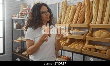 Donna che gusta croissant in una panetteria circondata da pane e dolci freschi, indossa occhiali e camicia bianca, mostrando felicità e assaporando la mamma Foto Stock