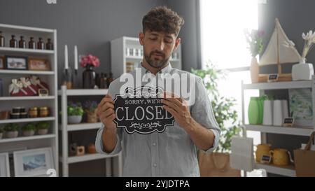 Un giovane con un cartello chiuso in un negozio di arredamento con scaffali di oggetti decorativi sullo sfondo che suggeriscono che il negozio non è aperto Foto Stock