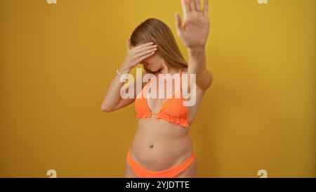 Donna che indossa un bikini arancione su sfondo giallo con la mano sollevata davanti al viso, che sembra proteggere gli occhi e bloccare la c Foto Stock