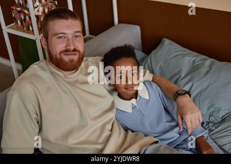 Ritratto di un bambino sorridente di colore che guarda la macchina fotografica con un felice padre adottivo mentre riposa insieme seduto sul divano nella camera dei bambini, copia spazio Foto Stock